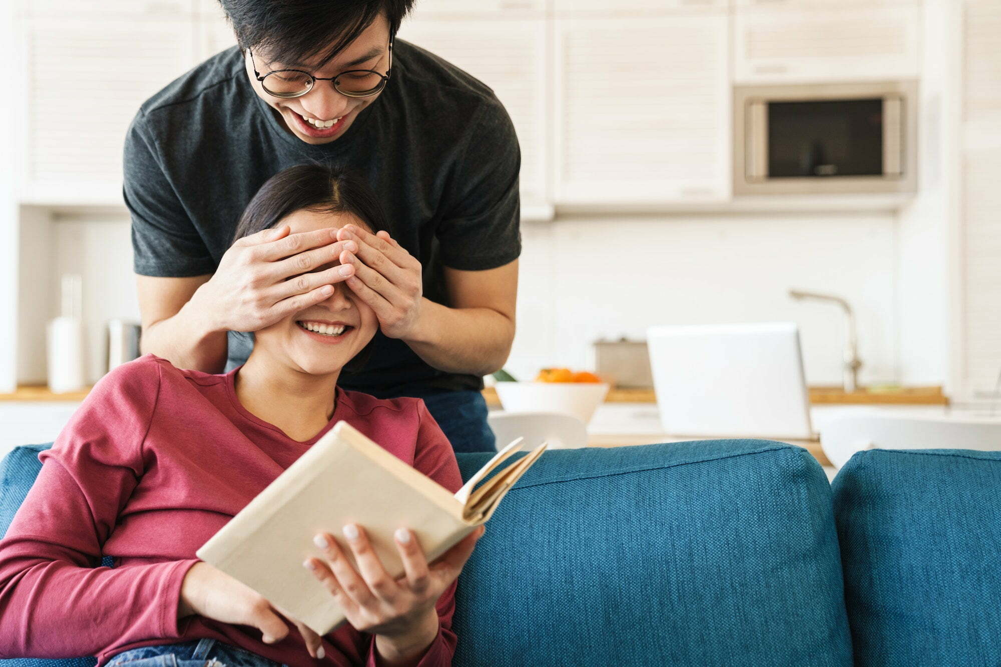 Photo of asian man covering girlfriend's eyes while she reading book