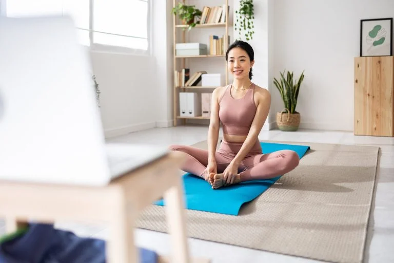 Young woman sitting on a yoga mat following an online fitness class on laptop